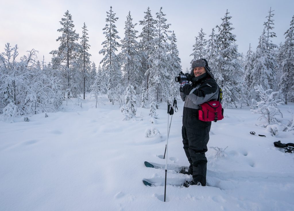 Mies talvisessa metsässä liukulumikenkien ja kameran kanssa